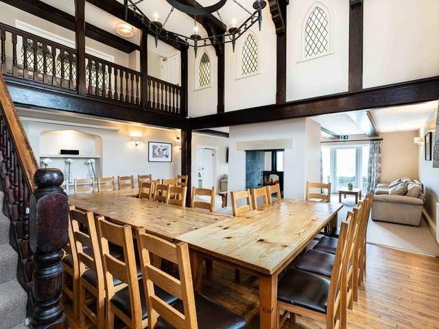 A room with numerous chairs around dining tables, with dark timber staircase to the left and balustrade above.