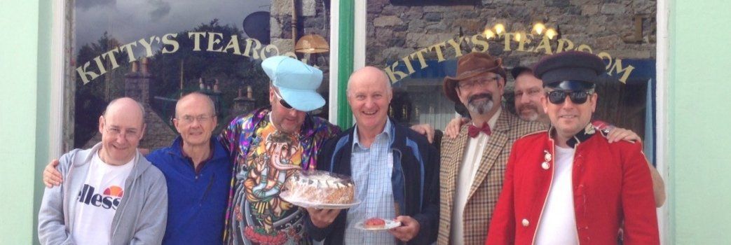 Photo of 7 men, some in fancy dress, with a cake in front of a tea shop window
