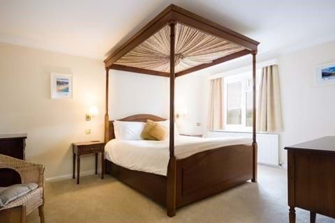 A wooden four-poster bed in the middle of a white-painted room with a window beyond